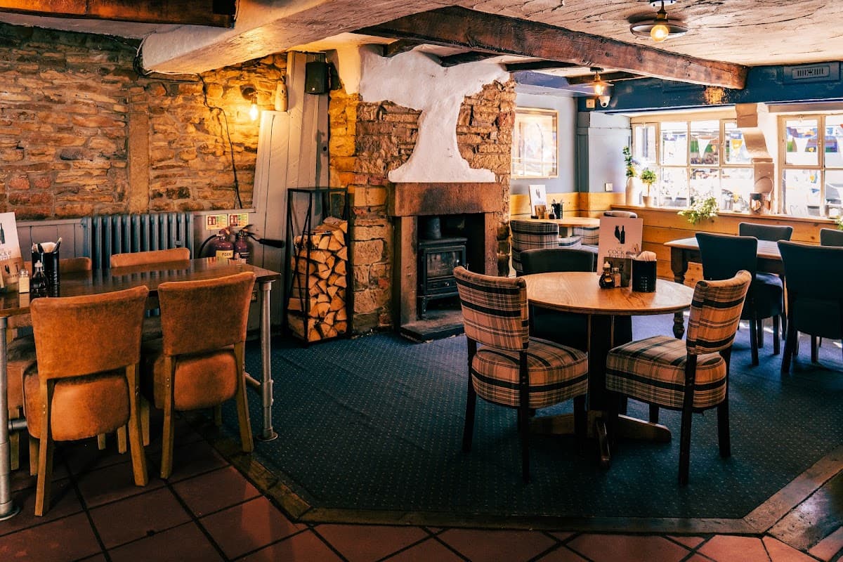 Cozy restaurant interior with stone walls, wooden beams, a fireplace, and tables set with chairs in warm tones.