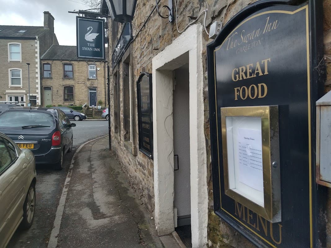 The Swan Inn sign, stone exterior, menu display, and parked cars along a quiet street in Carleton-in-Craven.
