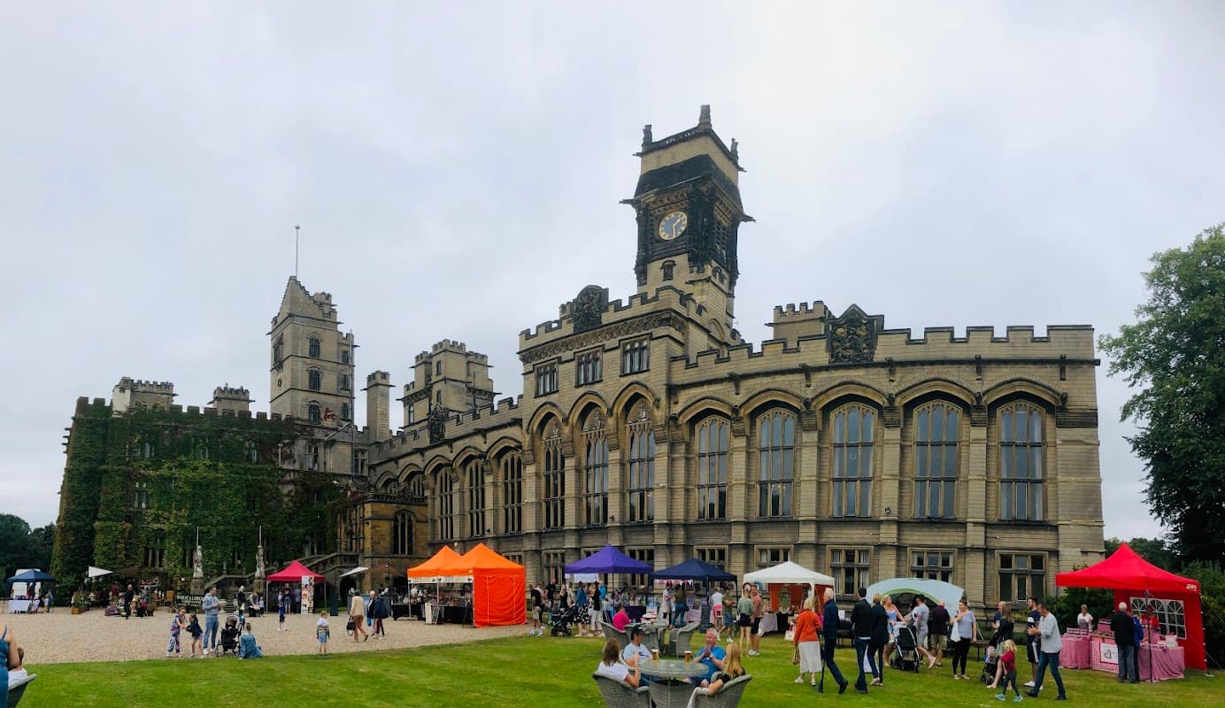 Historic building with clock tower, surrounded by green lawn and people at market stalls under colorful tents.