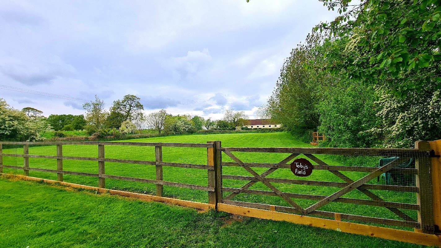Green field enclosed by a wooden fence, with trees and a building in the background under a cloudy sky.