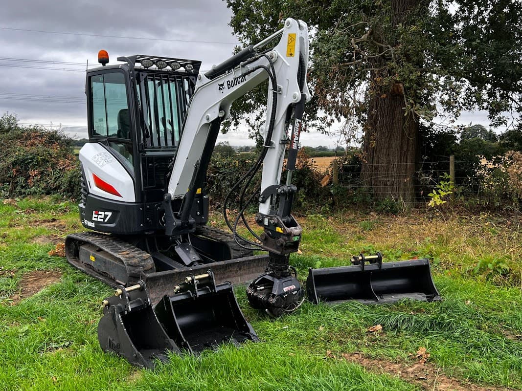 Excavator parked on grass with two attachments, surrounded by trees and open fields in North Yorkshire.