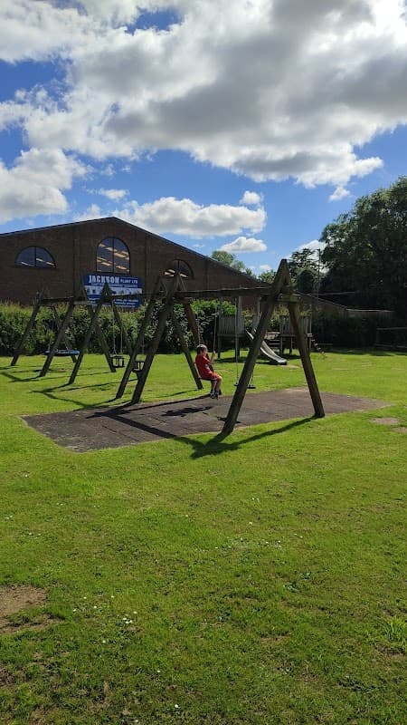 A playground with swings on grass, a child sitting on a swing, and a building in the background under a cloudy sky.