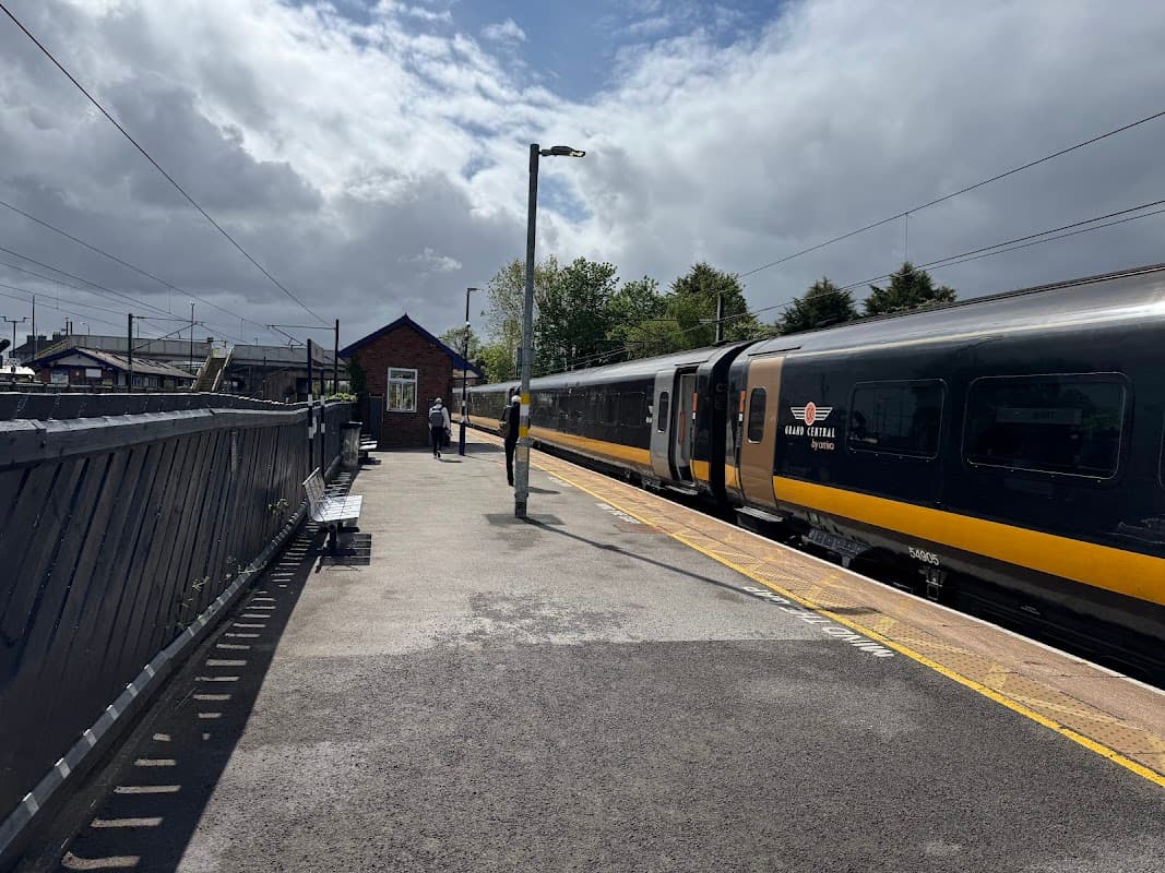Carlton Miniott Railway Station platform with a train, a passenger, and a station building under a partly cloudy sky.