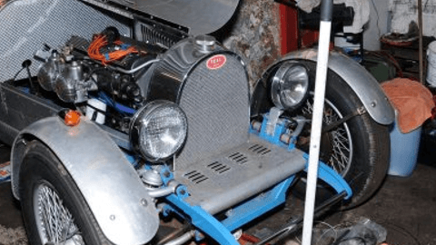 Classic car with exposed engine and chrome details, parked inside a garage with tools and equipment nearby.