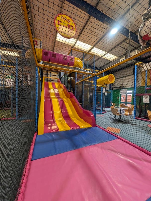 Brightly colored indoor play area featuring a large pink and yellow slide, climbing structures, and seating in the background.