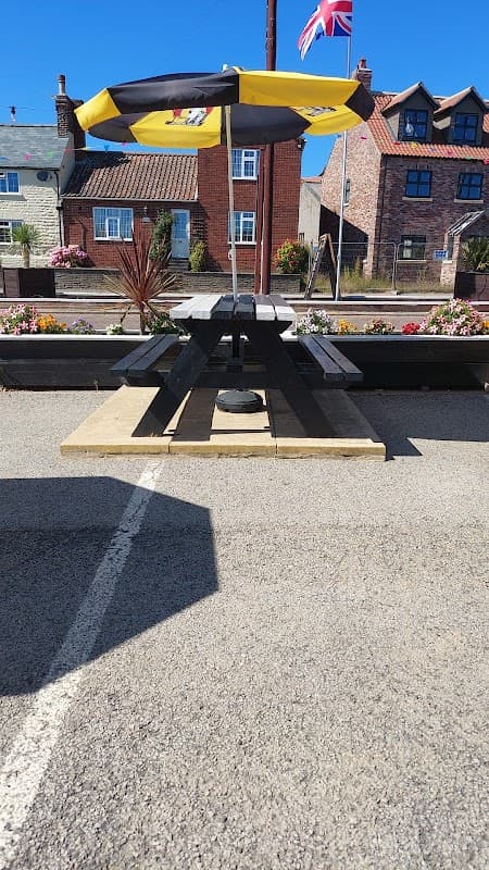 Black picnic table with a yellow and black umbrella, surrounded by flowers and buildings under a clear blue sky.