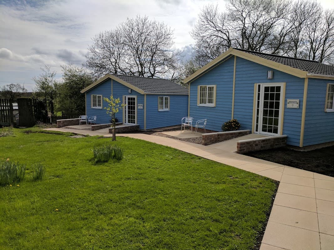 Two blue wooden cottages with white trim, surrounded by green grass and trees, featuring small patios and a pathway.