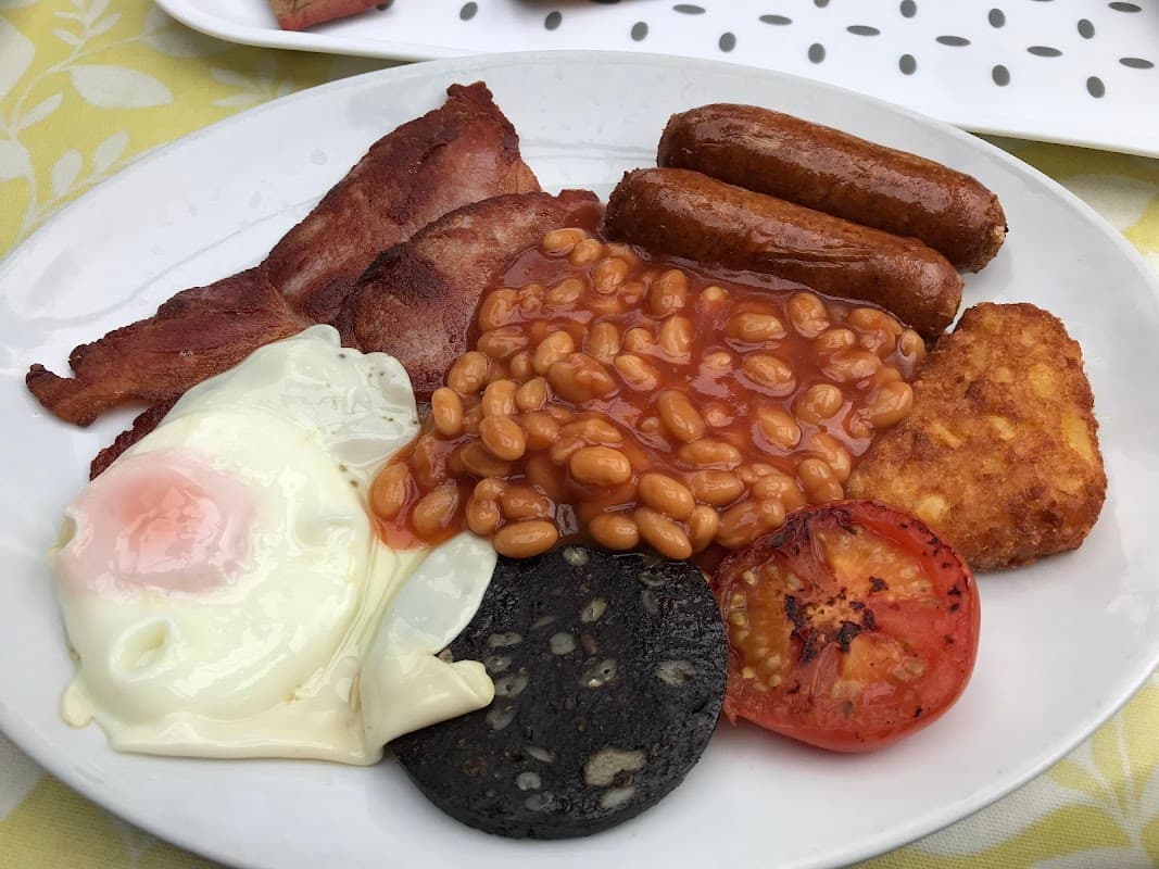 A plate featuring a full English breakfast: bacon, sausages, baked beans, fried egg, hash brown, black pudding, and tomato.