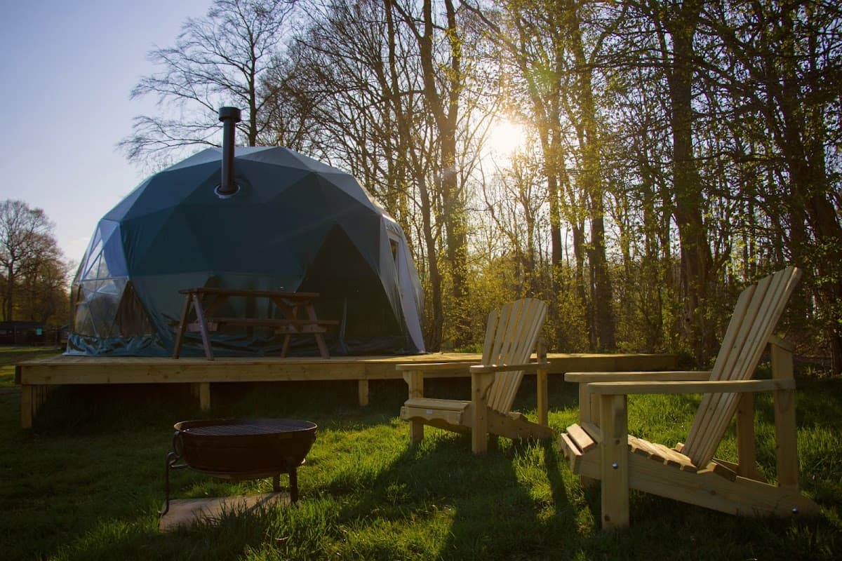 A geodesic dome structure with a chimney, surrounded by trees, wooden chairs, and a fire pit in a grassy area.