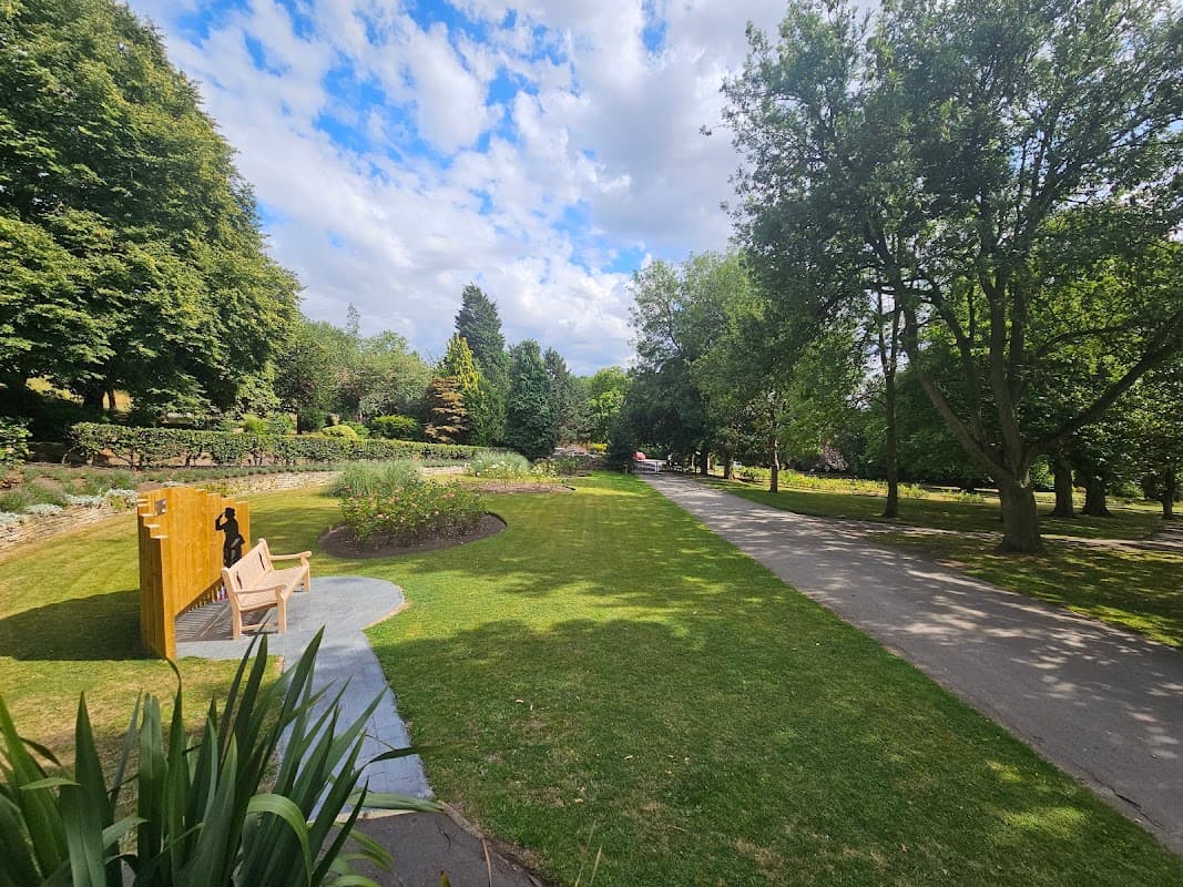 Lush green park with trees, a pathway, and a wooden bench near colorful flowerbeds under a partly cloudy sky.