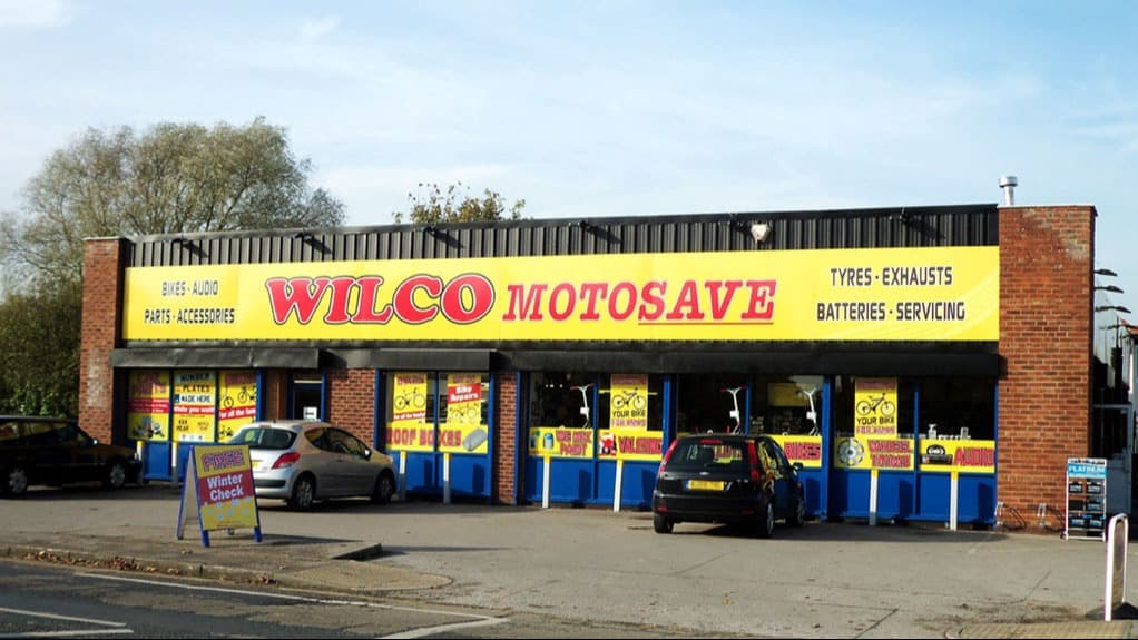 Bright yellow and red signage for Wilco Motosave, featuring tyres, exhausts, and batteries, with parked cars outside.