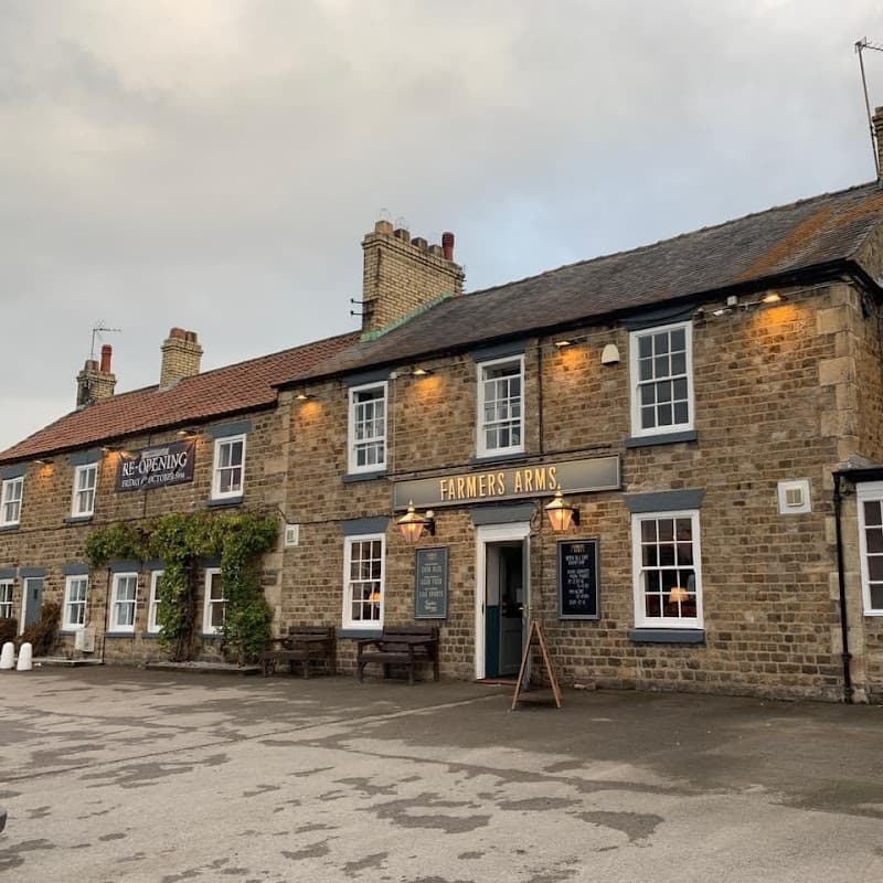 Historic stone building with a sign reading "Farmers Arms," outdoor seating, and a cloudy sky in Catterick Bridge.