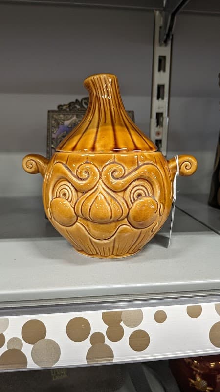 Brown ceramic pot with a whimsical face design, featuring a rounded body and a narrow neck, displayed on a shelf.