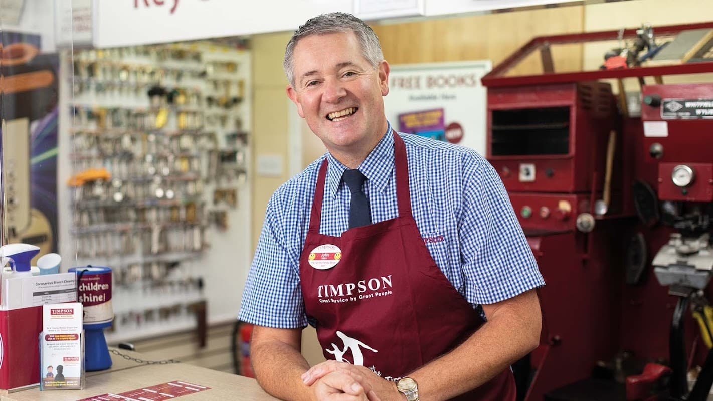 Smiling man in a red apron at a service counter with key cutting equipment and promotional materials in the background.