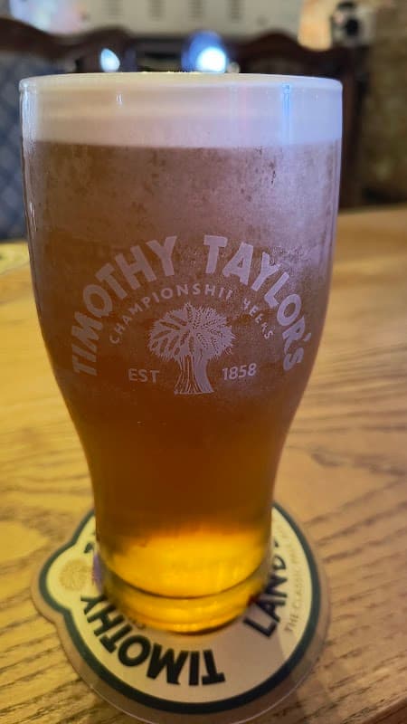 A pint of Timothy Taylor's beer on a wooden table with a coaster beneath it.
