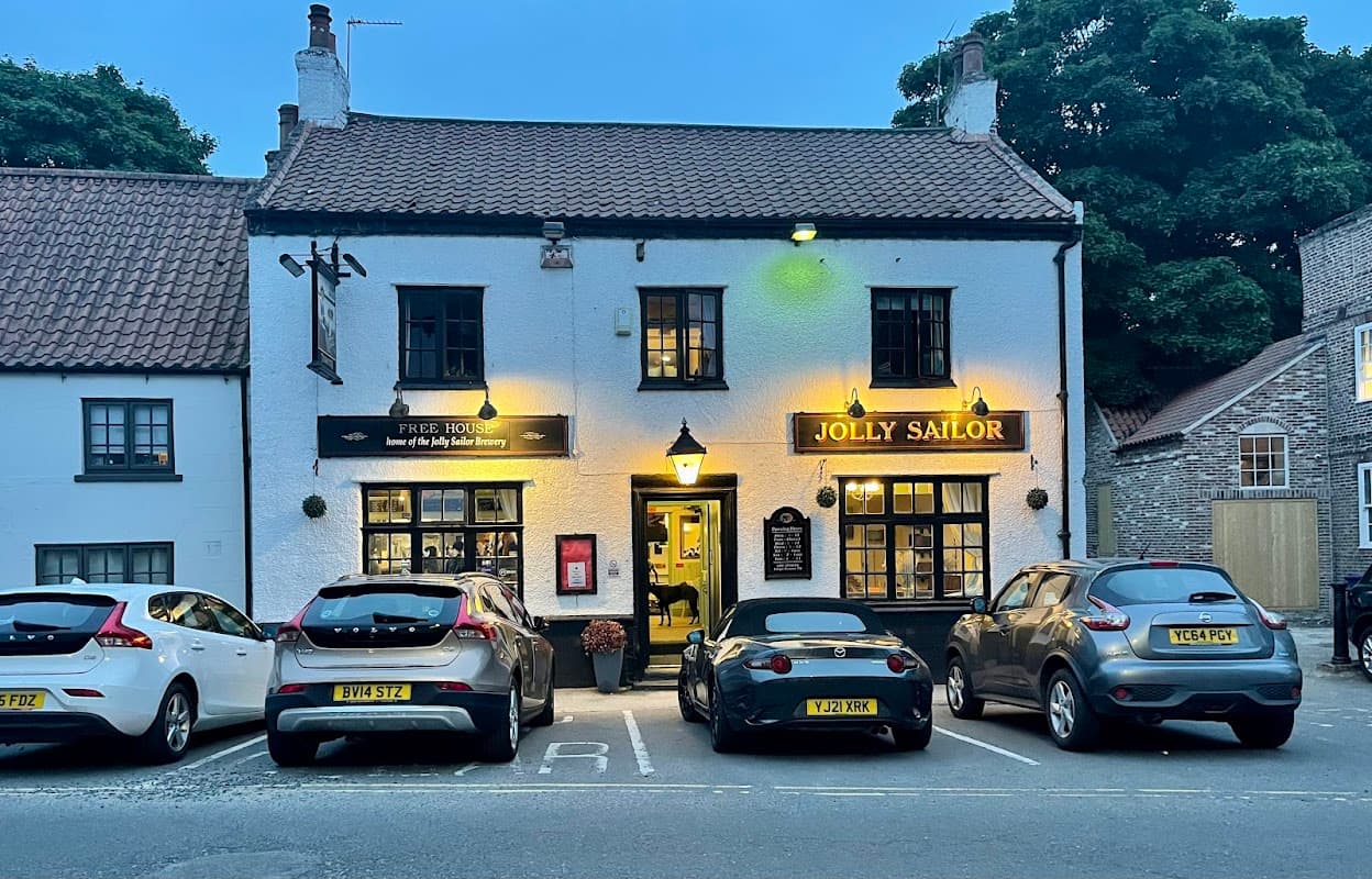 The Jolly Sailor Inn with a white exterior, warm lights, and parked cars in front, set in a quaint Yorkshire village.