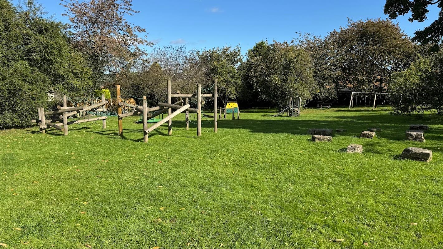 Playground equipment with climbing structures and swings in a grassy area surrounded by trees under a clear blue sky.