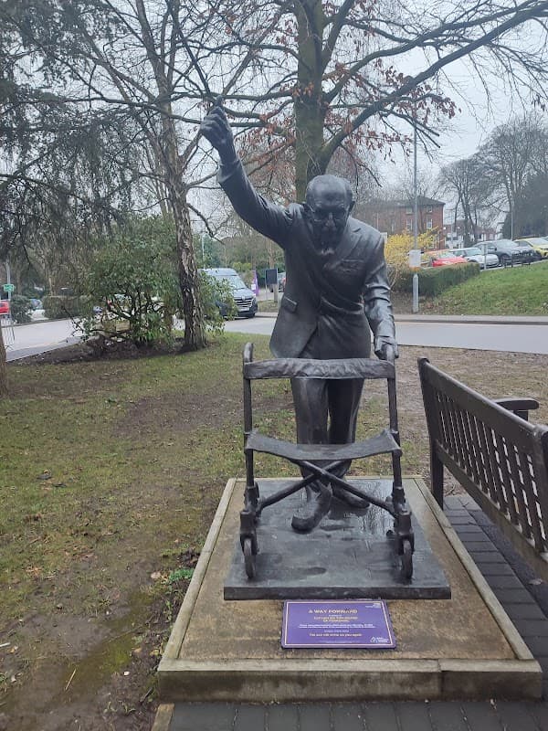 Bronze statue of a man raising his hand, beside a wheeled walker, set in a green area with trees and parked cars.