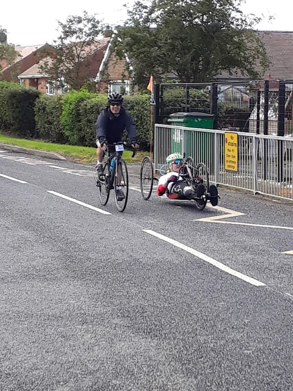 Cyclists on a road near Chapel Haddlesey Church of England Primary School, with greenery and a school sign in view.