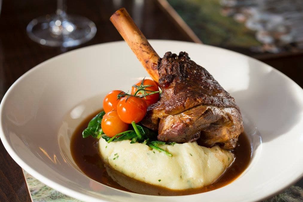 A plated dish featuring a lamb shank, cherry tomatoes, spinach, and creamy mashed potatoes in rich gravy.