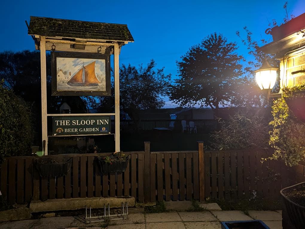 The Sloop Inn sign features a painting of a sailboat, surrounded by trees and a cozy beer garden at dusk.