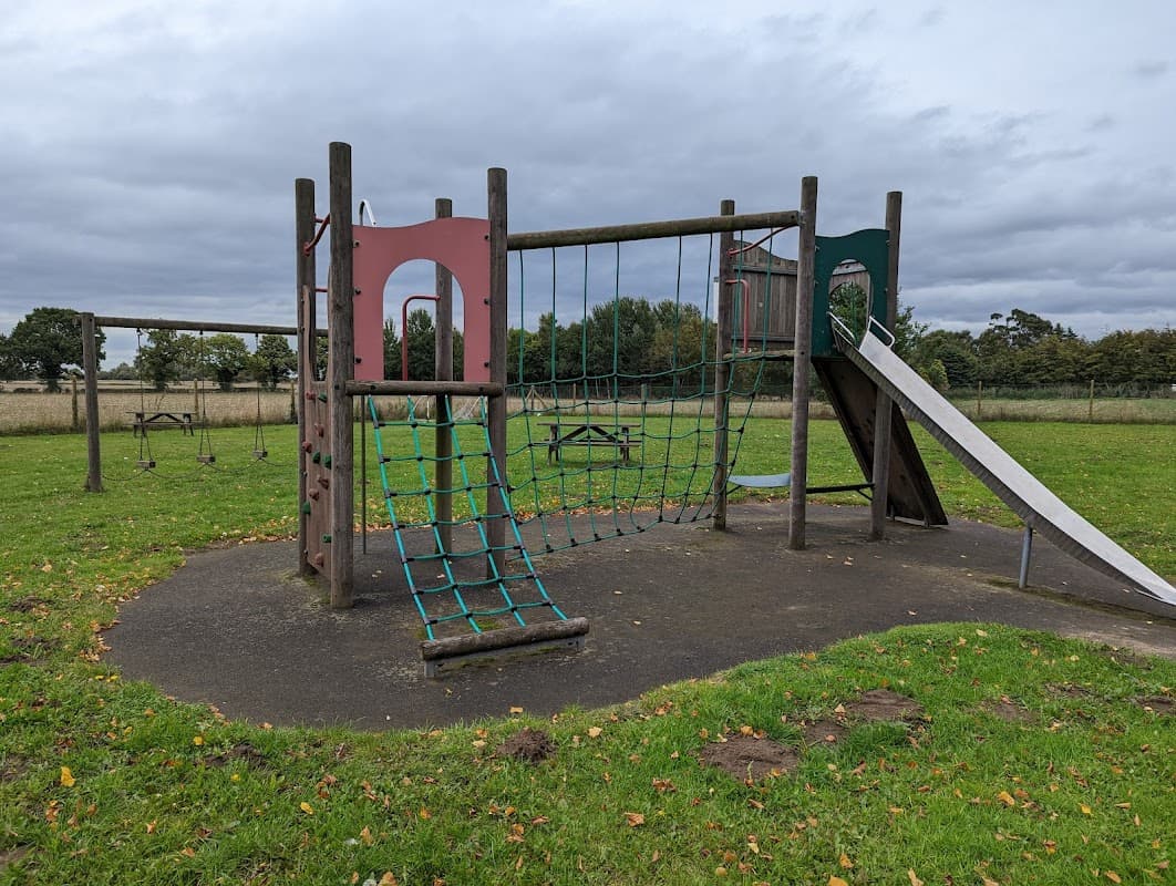 Playground equipment with a climbing net, slide, and swings on grassy field under a cloudy sky in West Haddlesey.