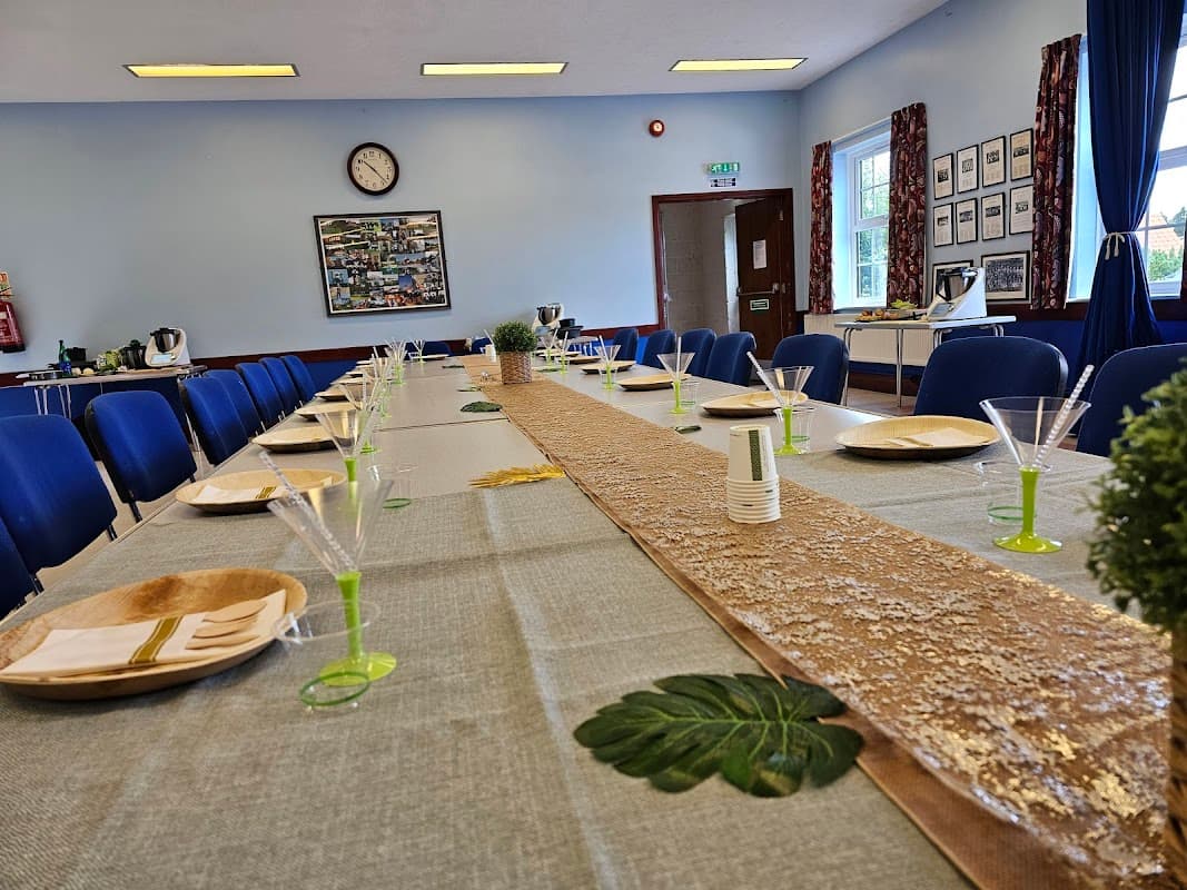 Long table set with green and gold decor, plates, glasses, and a clock on the wall in a bright, airy hall.