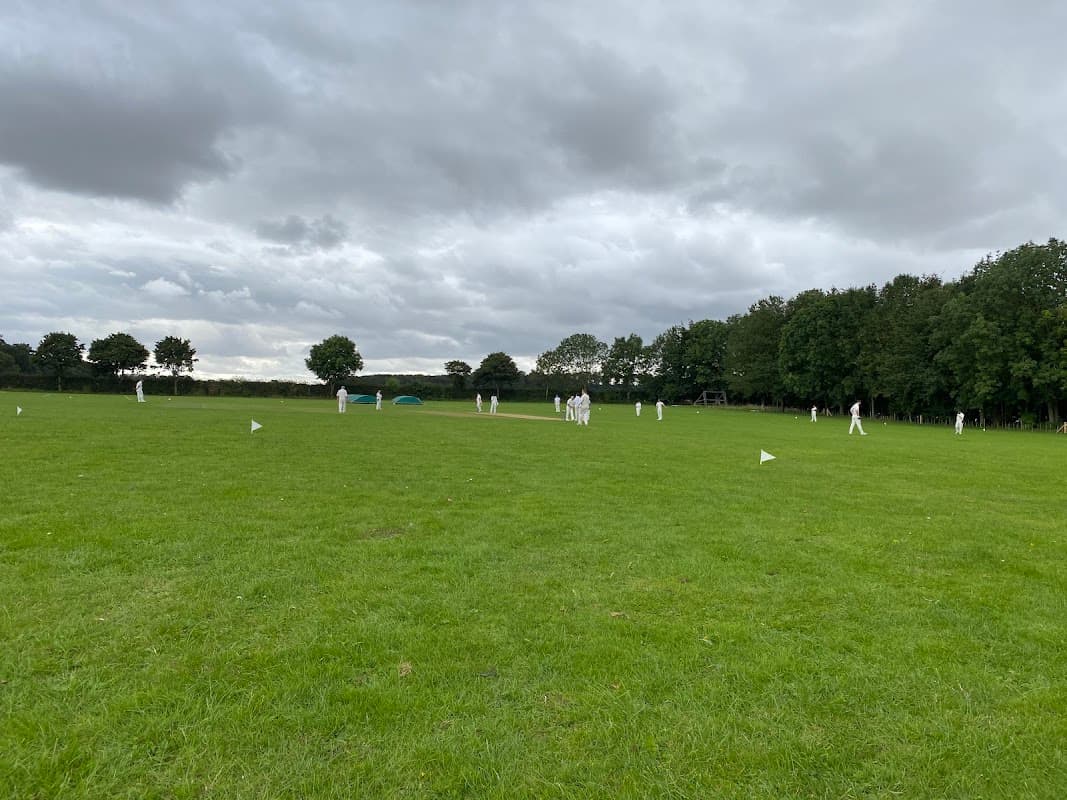 Cricket players in white uniforms on a lush green field under a cloudy sky, surrounded by trees.