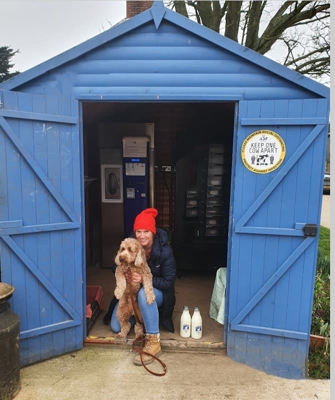 Blue shed entrance with a woman in a red hat holding a dog, surrounded by milk bottles and greenery.