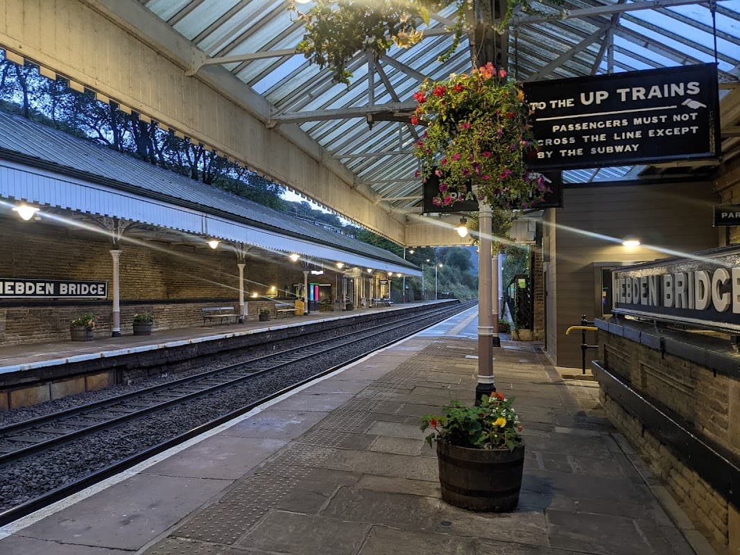 Hebden Bridge bus stop with a covered platform, flower baskets, and a sign directing to up trains. Quiet, early evening scene.