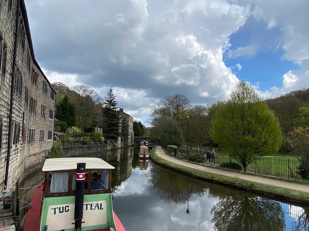 Canal scene with boats, stone buildings, trees, and a pathway alongside water under a partly cloudy sky.