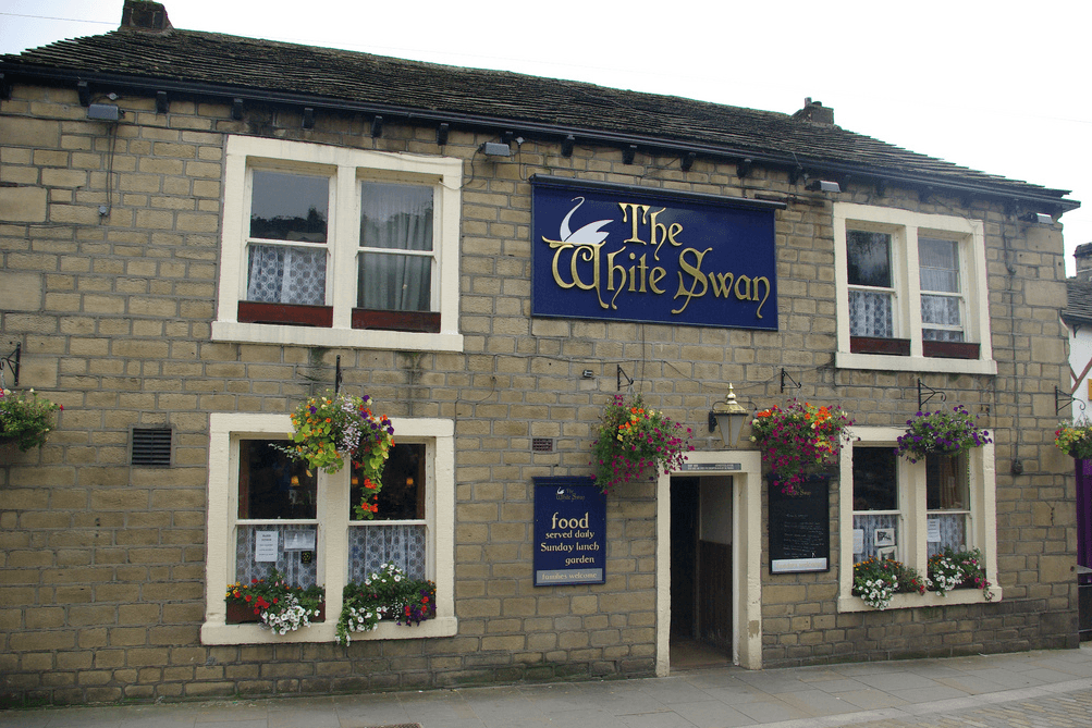 Stone building with a blue sign reading "The White Swan," adorned with colorful flower baskets and white-framed windows.