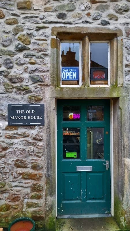 Green door with a sign reading "OPEN" and a plaque that says "THE OLD MANOR HOUSE," surrounded by stone walls.