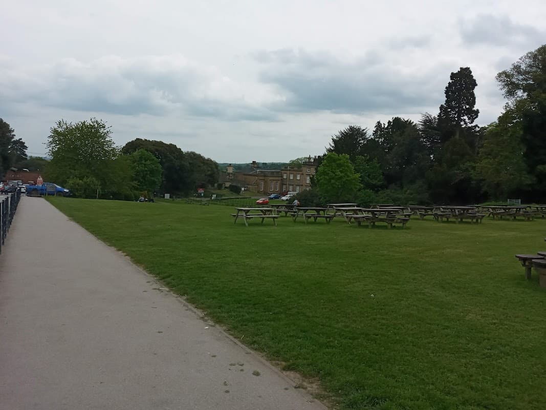 Spacious car park with picnic tables, green grass, trees, and a distant view of a building under a cloudy sky.