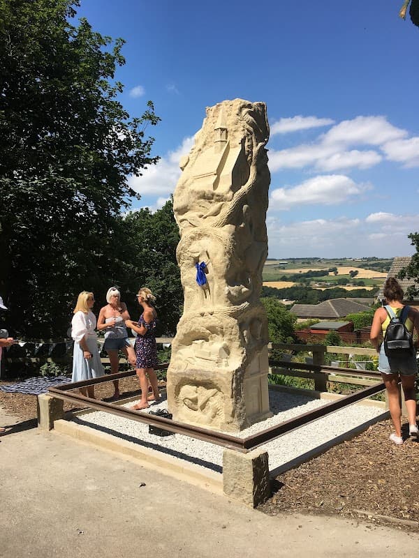 A tall stone sculpture surrounded by visitors, with rolling hills and a blue sky in the background.