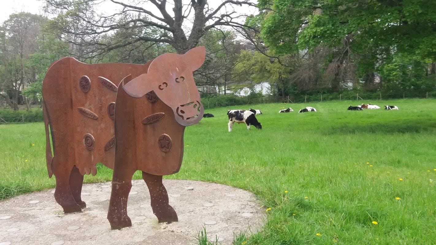 A metal cow sculpture in the foreground with grazing cows in a green field and trees in the background.