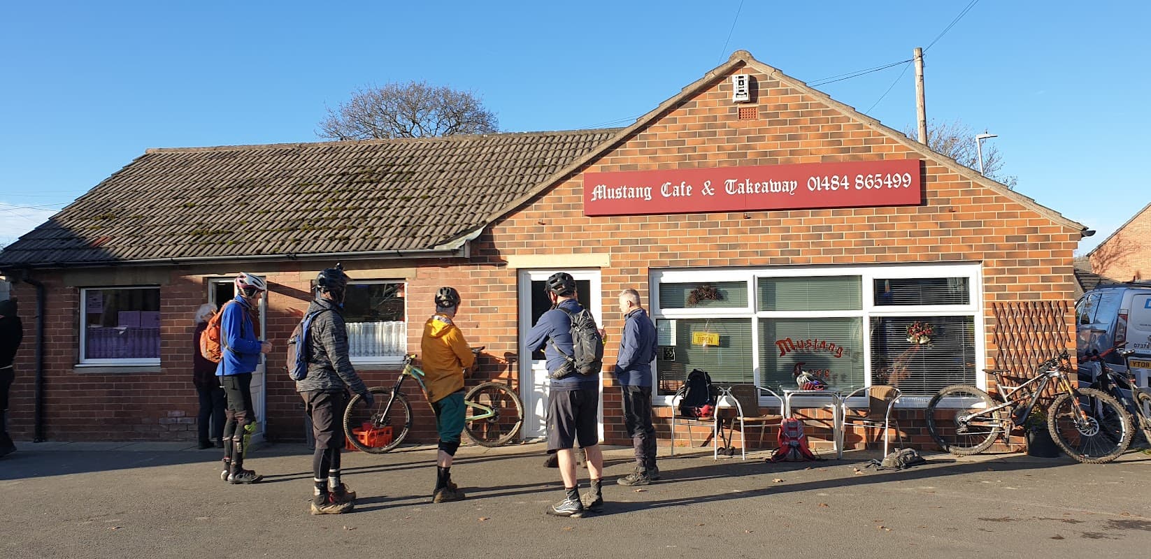 Customers queue outside Mustang Cafe & Takeaway, a brick building with a sign, bicycles parked nearby, on a sunny day.