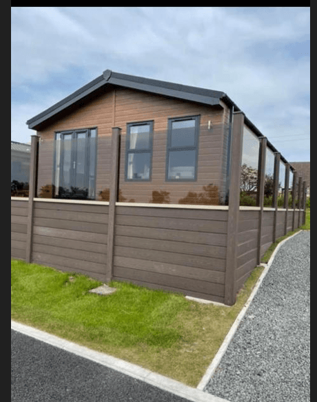 Modern holiday cabin with large windows, surrounded by a wooden fence and green grass, set against a cloudy sky.