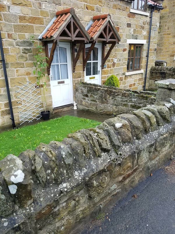Two cottage doors with wooden canopies, surrounded by a stone wall and a small green lawn in Cloughton, Yorkshire.
