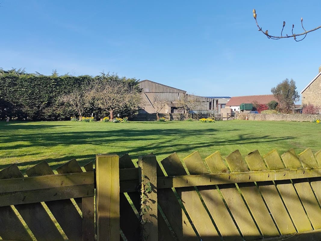 A green field with a wooden fence, surrounded by trees and buildings under a clear blue sky at Colburn Grange Farm.