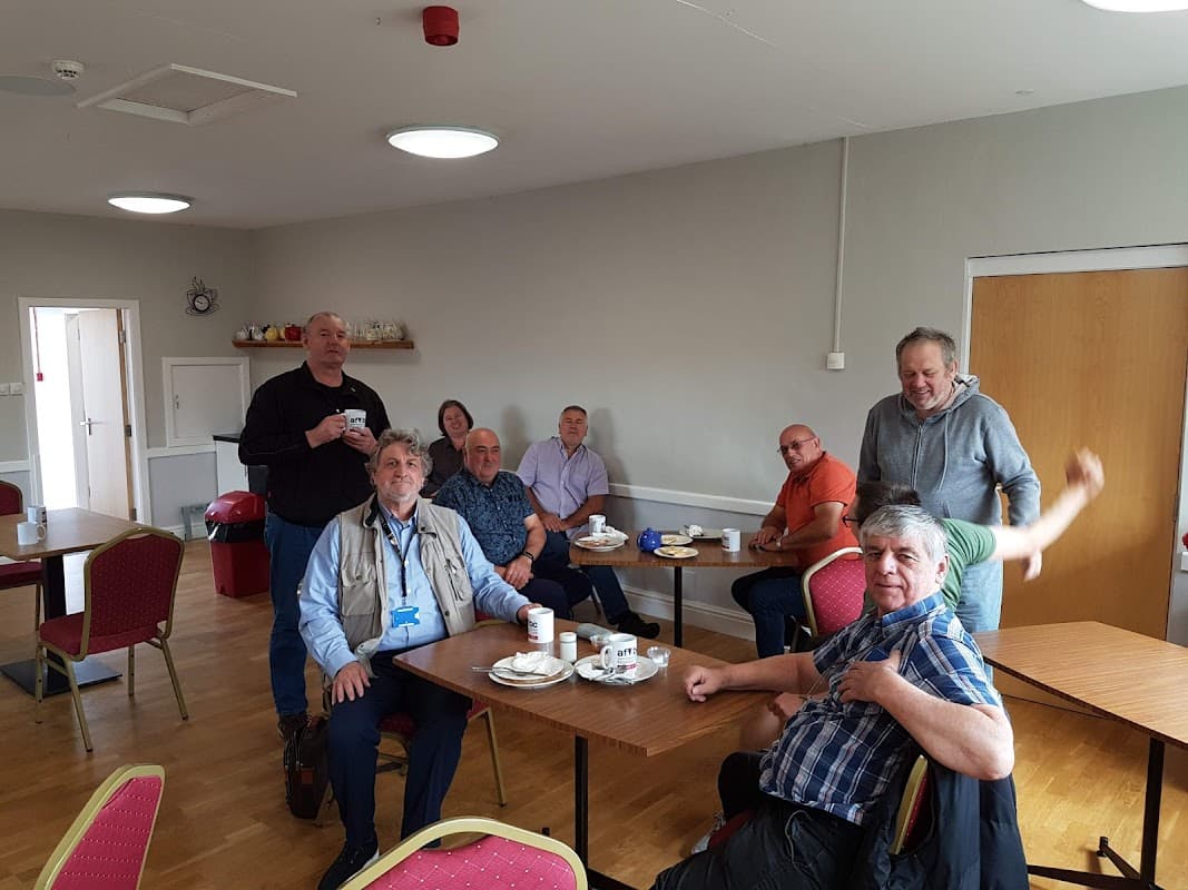 Group of people seated around tables in a community hall, enjoying drinks and snacks. Bright, welcoming interior.