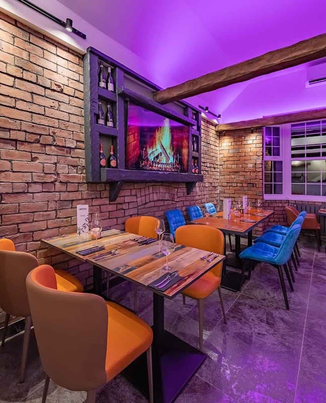 Cozy restaurant with colorful chairs, wooden tables, a brick wall, and a fireplace display under purple lighting.
