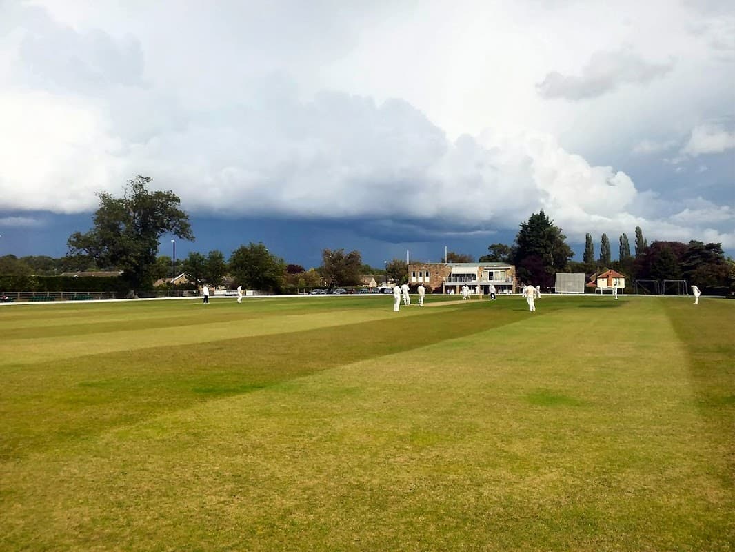 Cricket match in progress on a green field with cloudy skies and trees in the background at Collingham & Linton Cricket Club.