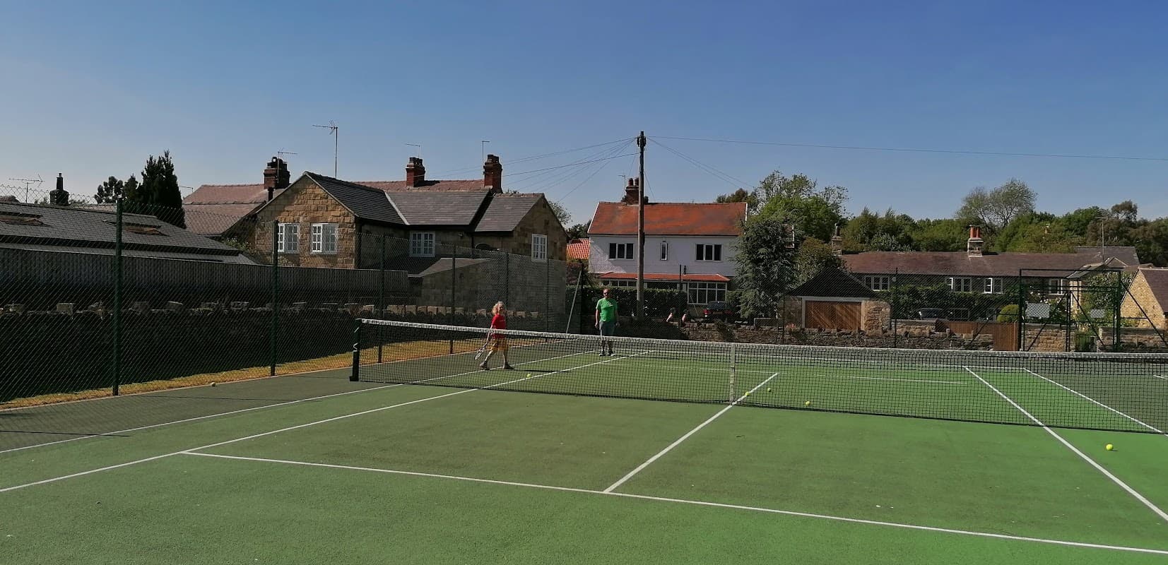 Tennis court with two players, surrounded by houses and trees under a clear blue sky in Collingham, Yorkshire.