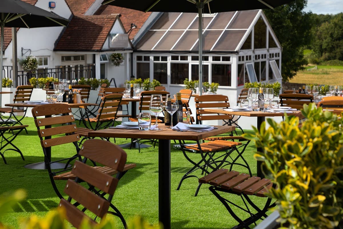 Outdoor dining area with wooden tables and chairs, surrounded by greenery and a glass conservatory in the background.