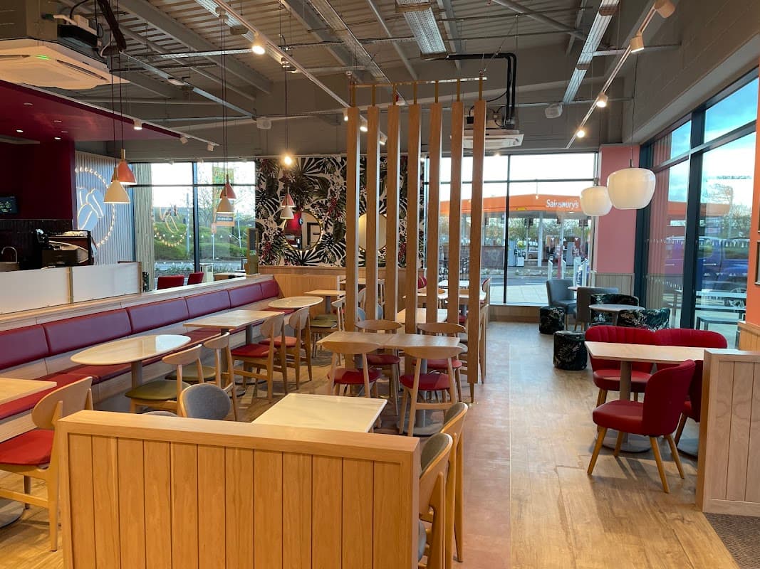 Modern coffee shop interior with wooden furniture, red seating, and large windows showing a drive-thru sign outside.