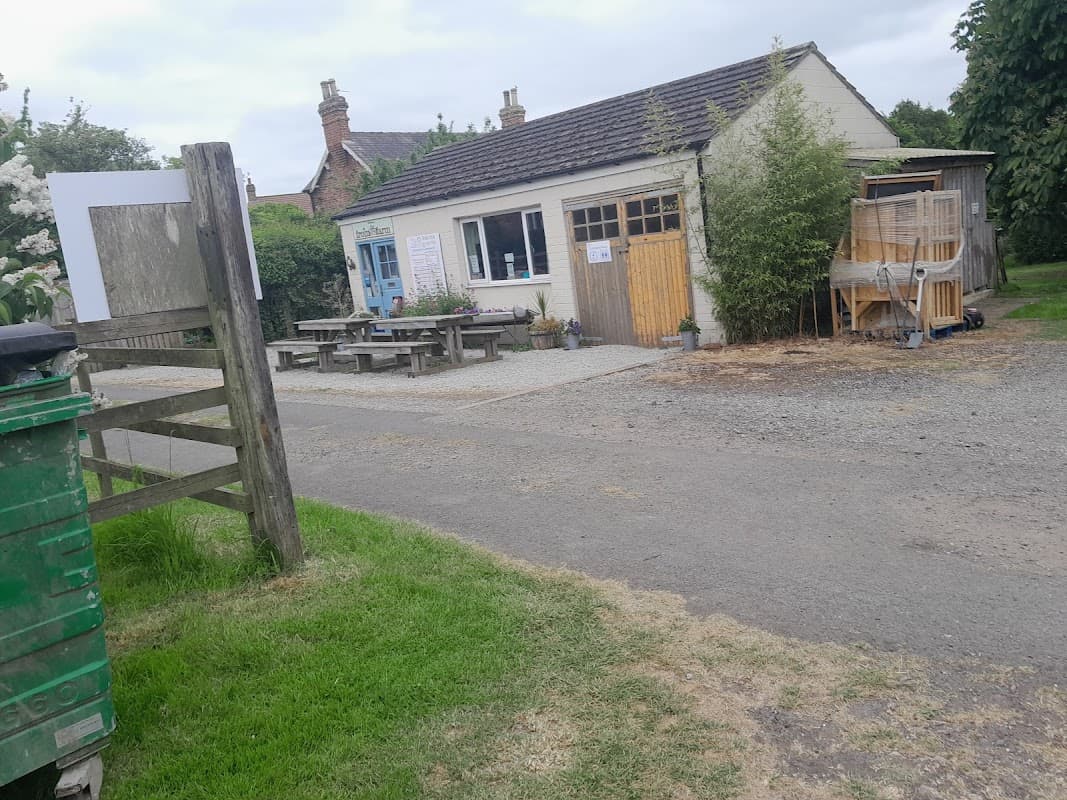 Campsite entrance with a building, picnic tables, green bin, and gravel path surrounded by trees and greenery.