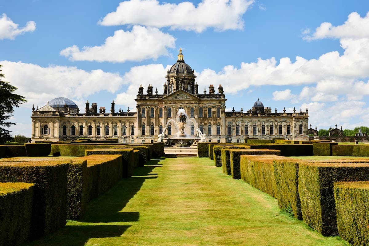 Grand Castle Howard with manicured hedges, a fountain, and a blue sky dotted with clouds in Coneysthorpe, Yorkshire.