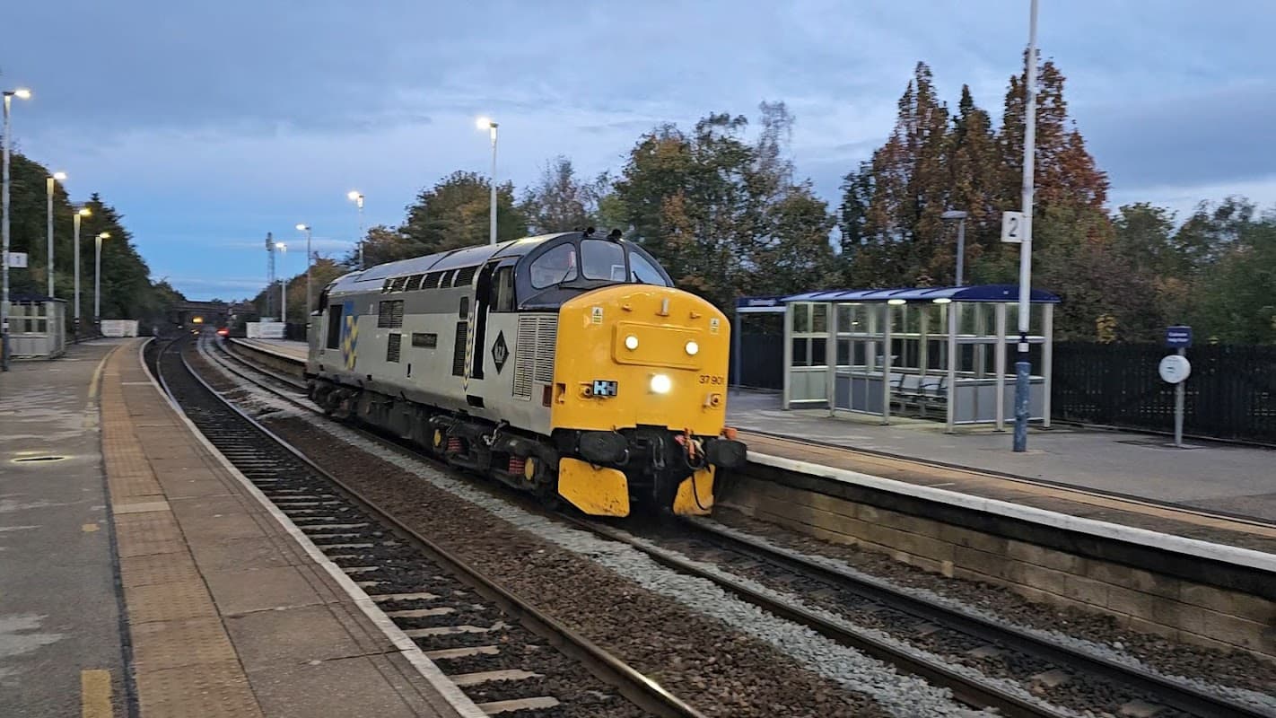 A train approaches a bus stop at Conisbrough station, with trees and a cloudy sky in the background.
