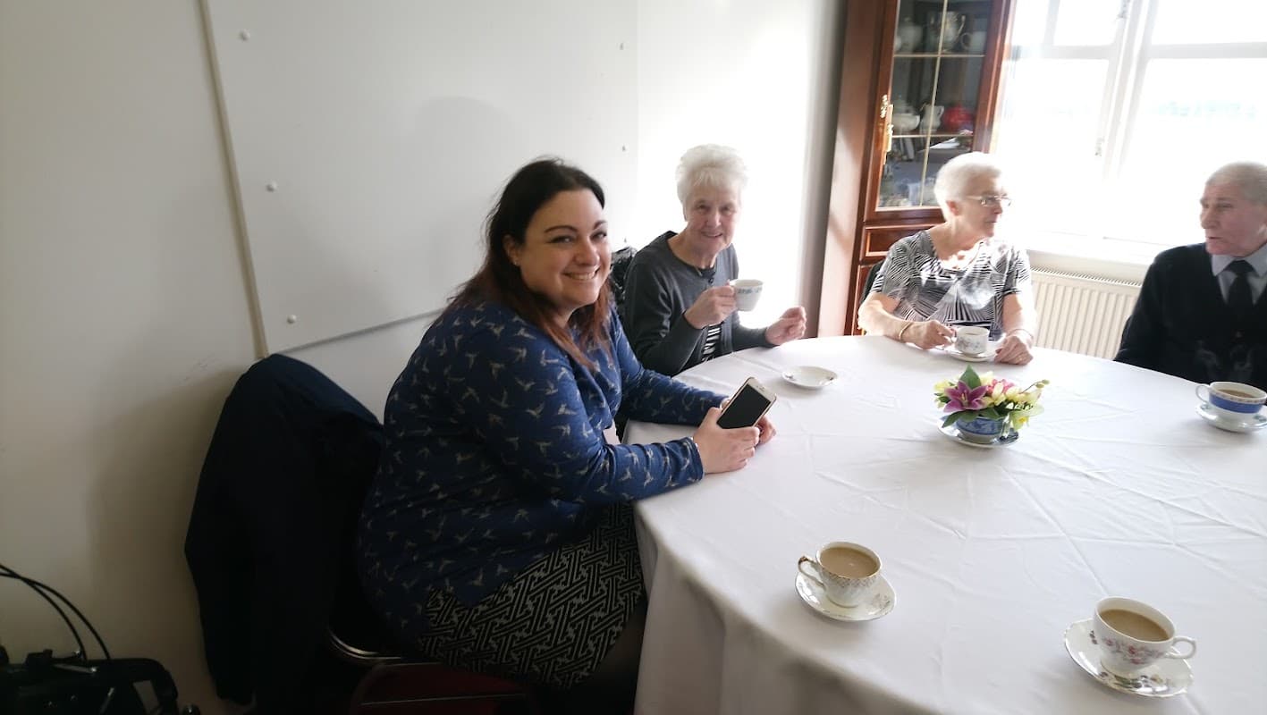 A group of four people enjoying tea at a round table in a bright room, with a floral centerpiece.